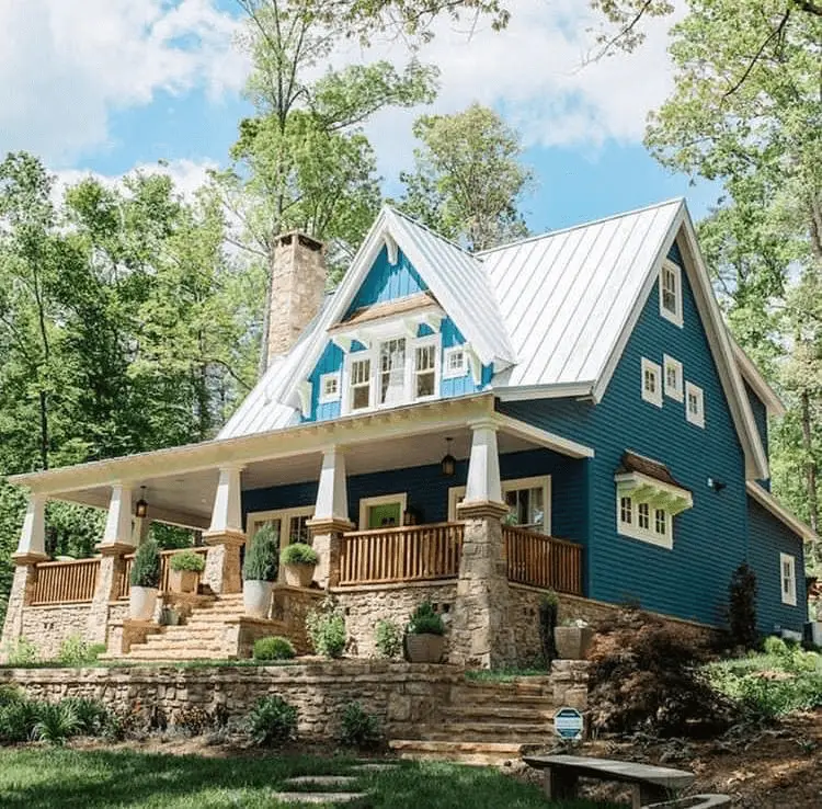 Design & Build - Cloudland Station: Striking teal cottage with white metal roof, stone pillars, and wooden porch, nestled among vibrant forest greenery.